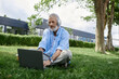 © Bliss - A mature gay man with tattoos and a grey beard sits on the grass, working on his laptop.