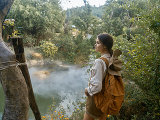 A woman with a brown backpack standing near a body of water with steam coming out of it