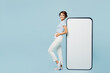 © ViDi Studio - Full body side view young woman wear striped t-shirt casual clothes stand near big huge blank screen mobile cell phone smartphone with mockup area isolated on plain blue background. Lifestyle concept
