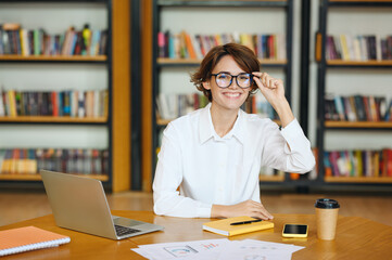 Wall Mural - Young smiling happy cheerful fun successful employee business woman wearing white shirt casual clothes touching glasses look camera sit work at office desk with pc laptop. Achievement career concept.