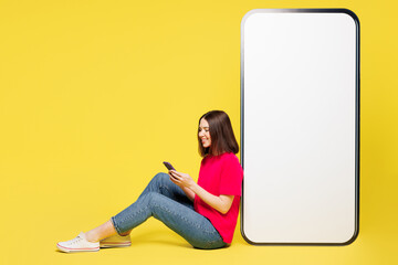 Wall Mural - Full body side view young woman wears pink t-shirt casual clothes sits big huge blank screen mobile cell phone with area use smartphone isolated on plain yellow orange background. Lifestyle concept.