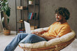 © ViDi Studio - Side view young Indian IT man wear orange casual clothes hold use work on laptop pc computer sits in armchair stay at home hotel flat rest relax spend free spare time in living room. Lounge concept.