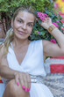 © Tymon - Elegant Blonde Woman in a White Sleeveless Dress Posing Amidst Lush Greenery and Vibrant Pink Blooms