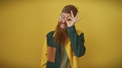 Canvas Print - Cheerful young redhead bloke with glasses, bearded and handsome, offering a cheeky grin while standing over isolated yellow background. fun 'ok' sign gesture with hand, looking through fingers.