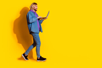 Full length photo of lovely young male hold netbook walking dressed stylish jeans garment isolated on yellow color background