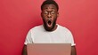 © AS Studio - genuine reaction of a men surprised by laptop news, isolated on a red color background with ample copy space, showcasing his wide-eyed expression and shocked demeanor