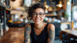 © ifoto - portrait of smiling woman with glasses and tattoos sitting at a table in a restaurant, the woman is wearing a black sleeveless top, she has short curly hair,