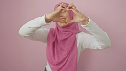 Poster - Cheerful young hispanic woman in hijab forms heart shape with hands, radiating love. a beautiful portrait against an isolated pink background, a romantic concept captured over.