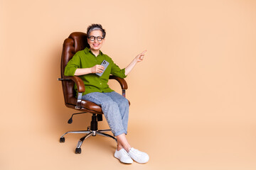 Poster - Photo portrait of lovely pensioner lady point empty space sit armchair dressed stylish green garment isolated on beige color background