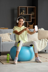 Poster - Sporty young African-American woman on fitball pointing at mobile phone in living room