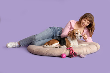 Wall Mural - Young woman with cute beagle dog and toys lying on pet bed against lilac background