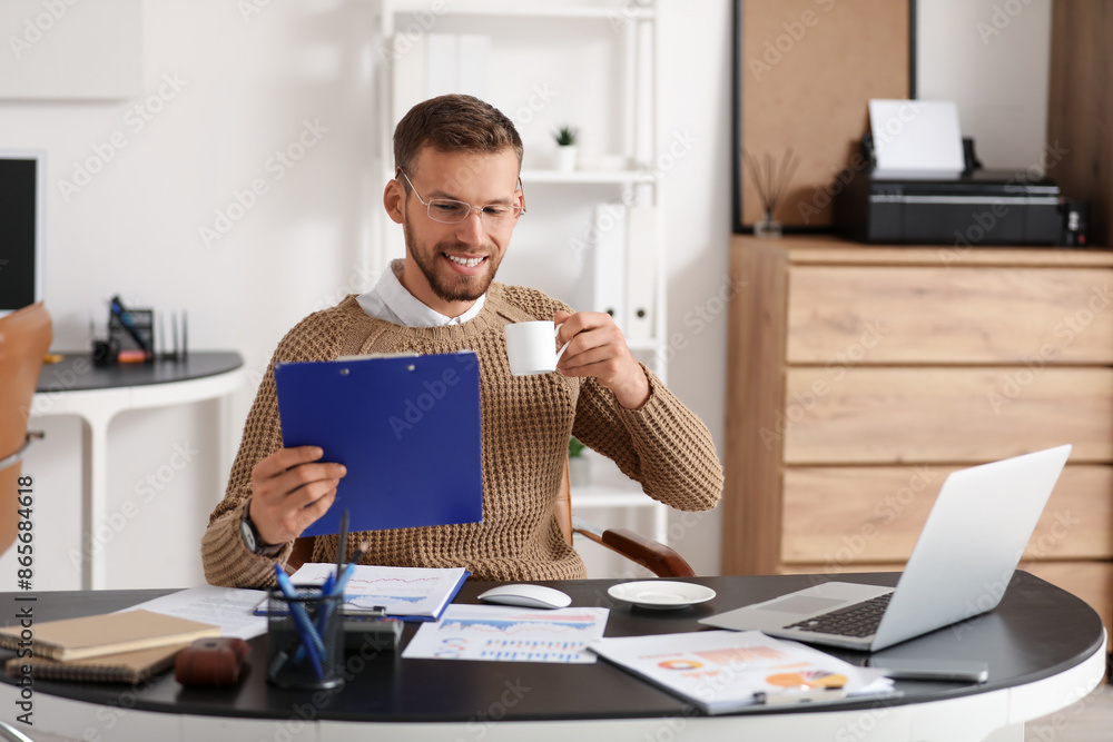 Young businessman in eyeglasses working with documents and drinking coffee at workplace in office