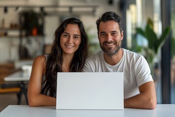 A couple sits at a table with a blank laptop screen in front of them, smiling at the camera.