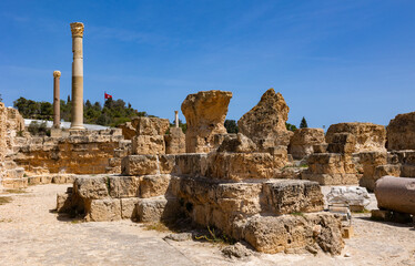 Wall Mural - Scenic view of ruins of Roman Baths of Antoninus at Tunisian archaeological site of Carthage, featuring remnants of tall Corinthian columns and stone structures under blue sky..