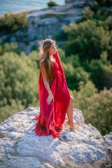 Wall Mural - Woman sunset sea red dress, back view a happy beautiful sensual woman in a red long dress posing on a rock high above the sea on sunset.