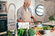 © luciano - Smiling bearded senior man holding frying pan with vegetables. People in the kitchen preparing dinner. An elderly man alone cooking at home