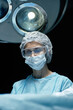 © Seventyfour - Vertical portrait of mature female surgeon wearing mask performing operation in blue light at hospital