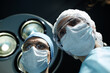 © Seventyfour - Low angle portrait of two surgeons wearing masks looking down under surgical lamp while performing operation