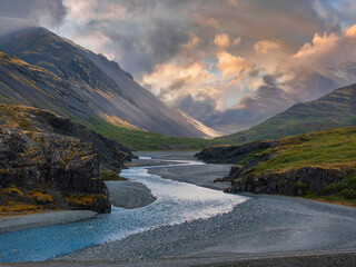 A light blue river winds through a dark gray mountain valley under a cloudy sky in Iceland. The image captures the stunning beauty of the Icelandic landscape.