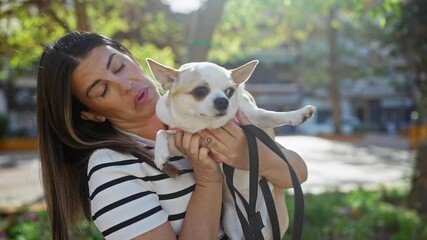 Wall Mural - Young woman holding a chihuahua in an urban park displaying humorous reaction with sunlight filtering through the trees.