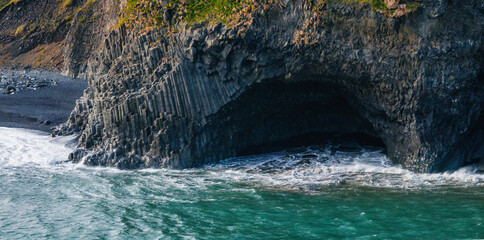 Dramatic aerial image of black volcanic rock formation with hexagonal columns and cave opening, located on the coast of Iceland with waves crashing against the rocks.