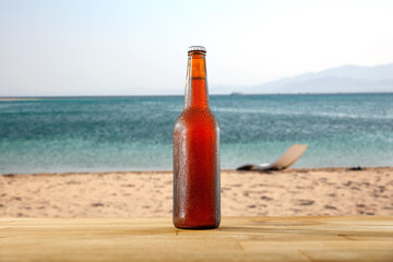 Wall Mural - Glass bottle of cold beer and ice on wooden plank on sandy shore of a beach with copy space