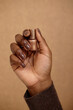 © Seventyfour - Vertical shot of Black woman holding golden nail polish for creating manicure. Dark brown nails pairing lacquer and skin tone of girl on beige background