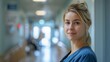© Helen-HD - Female nurse in a blue scrubs stands in a hospital hallway, smiling softly at the camera. The background is blurred, creating a sense of depth and privacy