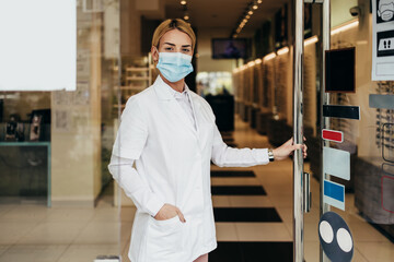 Wall Mural - Young and attractive female optometrist with face protective mask standing at open optical store doors and looking outside. She is confident and serious. Covid-19 open for business concept.