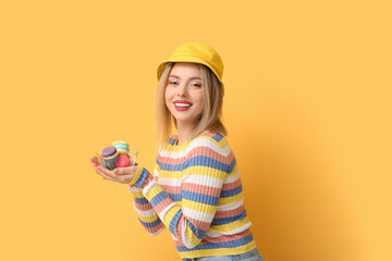 Poster - Happy young woman holding glass bowl with sweet macaroons on yellow background