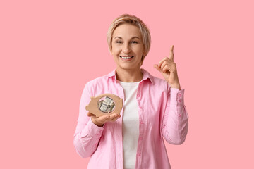 Poster - Happy mature woman with wooden piggy bank pointing at something on pink background