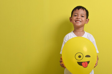 Tongue emoji. Cute Asian boy holding tongue out emoticon balloon isolated on yellow background. Teasing emoji with closed eyes