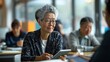 © nilanka - Senior adult woman wearing glasses seated in a classroom and use a tablet or mobile phone. Acticve diverse senior citiziens concept.