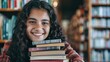 © chutikan - A young woman smiles with a pile of books on the background of a bookshelf in a library.