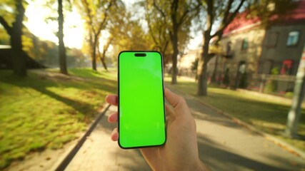 Young Caucasian man using mobile phone while walking through public park. Close-up male hand holding smartphone with green screen in vertical position and scrolling through social media feed outdoors