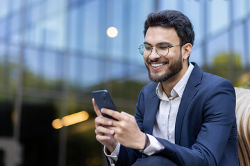 Smiling businessman in suit using smartphone outdoors. Confident professional checking messages through mobile phone with modern glass building. Concept of business, technology, communication