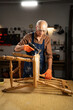 © Andrii Lysenko - Elderly man in a workshop using a drill with a special attachment to remove old varnish from a wooden chair. He is wearing protective goggles and orange gloves.