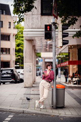 Wall Mural - Woman in casual outfit leaning against a pole, checking her phone while waiting at a red pedestrian light in an urban area.