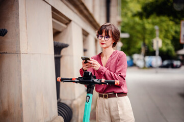 Wall Mural - Woman in casual attire standing on a sidewalk, using her phone next to a parked electric scooter.