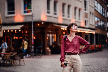 Canvas Print - Woman in casual attire stands confidently with her hand on her hip, holding her phone, in a bustling city street with cafes and lights.