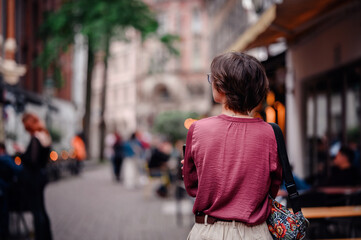 Wall Mural - Woman in casual attire with a colorful backpack walking through a bustling city street filled with people and cafes.