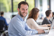 © pham - A man with a beard smiles at the camera while working on his laptop in a modern office setting