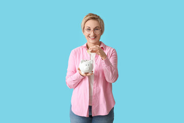 Wall Mural - Happy mature woman putting coins into piggy bank on blue background