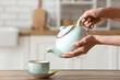 © Pixel-Shot - Woman pouring tea from teapot into cup on table in kitchen