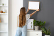 © Pixel-Shot - Young woman hanging frame on wall at home
