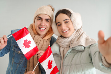 Canvas Print - Young couple in warm clothes with flags of Canada taking selfie on light background, closeup