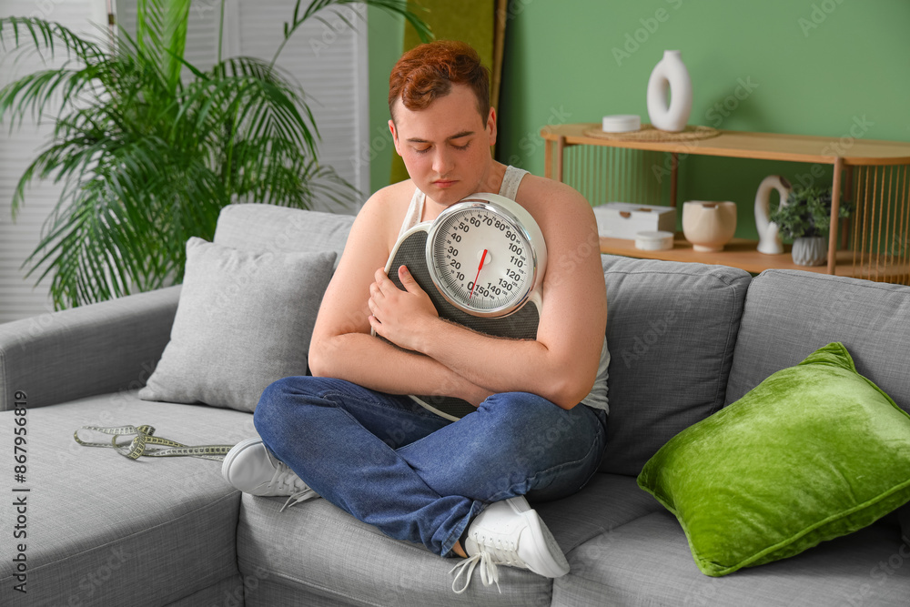 Young sad overweight man with scales sitting on sofa at home. Weight gain concept