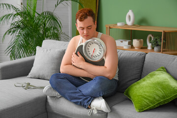 Sticker - Young sad overweight man with scales sitting on sofa at home. Weight gain concept