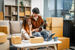 © NanSan - A young couple works from home, packing orders and managing their online store. They use technology and teamwork to run their business, smiling while fulfilling customer orders.