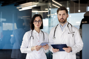 Wall Mural - Two doctors in white coats standing in a modern hospital setting. The male doctor holds a clipboard, and the female doctor uses a tablet. Projecting professionalism and medical expertise.